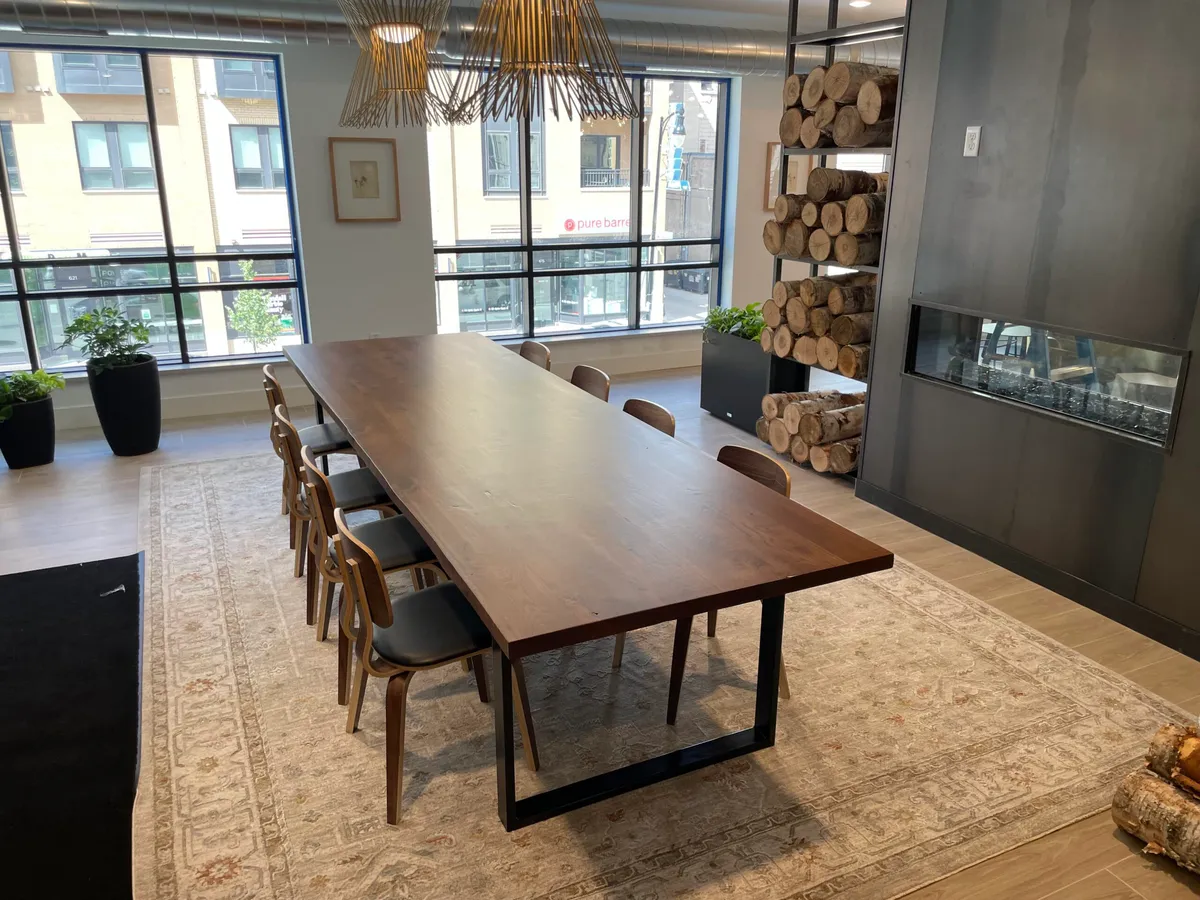 Large live-edge wood dining table with black steel legs, wooden chairs, and a stacked-log display beside a modern fireplace