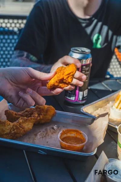 Hands holding a saucy fried chicken piece over a tray with dipping sauce and a canned drink