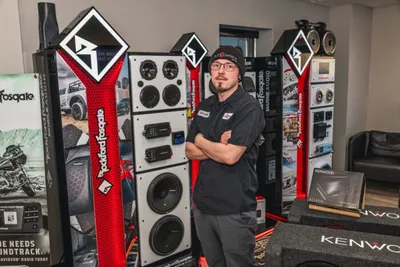 Man with crossed arms stands beside tall car audio display towers in a shop showroom