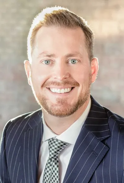 Smiling man with blue eyes and short red beard wearing a navy pinstripe suit and patterned tie