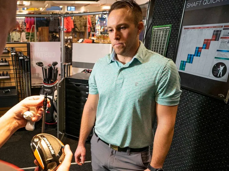 Man in striped polo examines a golf club head inside a sporting goods fitting studio