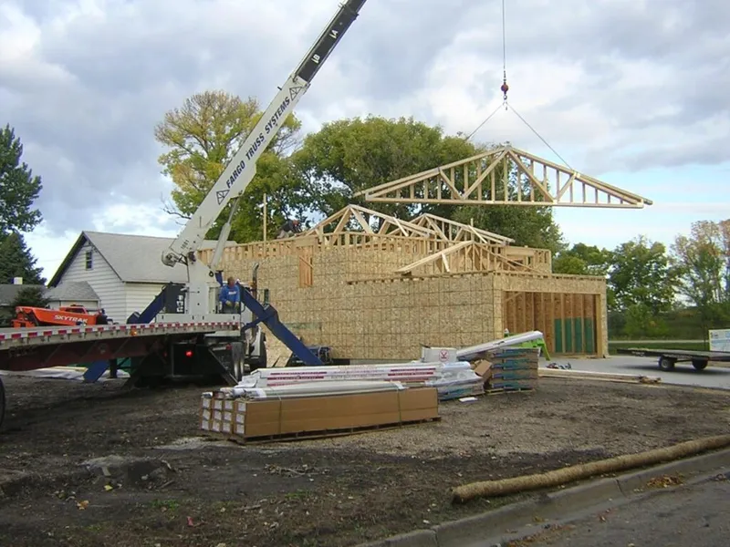 Crane lifting wooden roof trusses onto a partially framed house at an active construction site