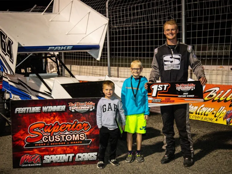 Sprint car race winner poses with two young boys holding 1st place and feature winner signs at a dirt track
