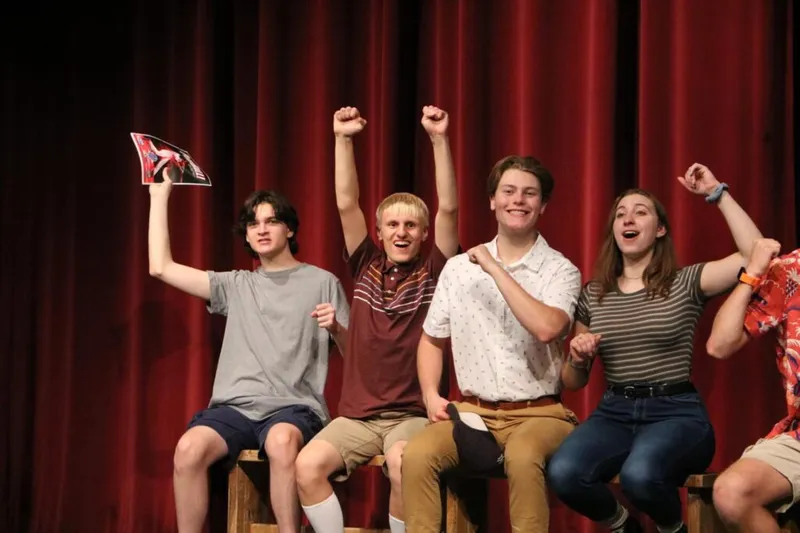 Four cheering teenagers sit on a stage bench in front of a red curtain