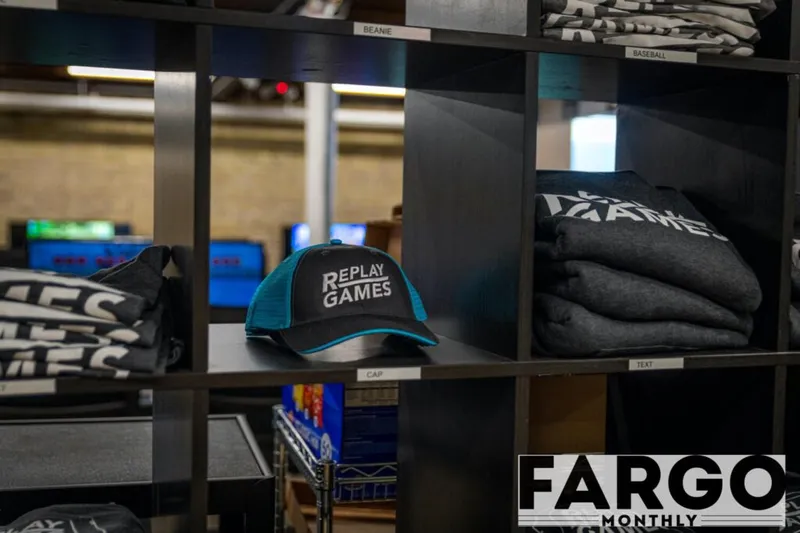 Black and teal Replay Games branded cap on a retail shelf beside folded branded shirts