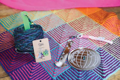 Colorful knitted blanket with geometric squares, a skein of dark yarn, stitch markers, and a small notions tin