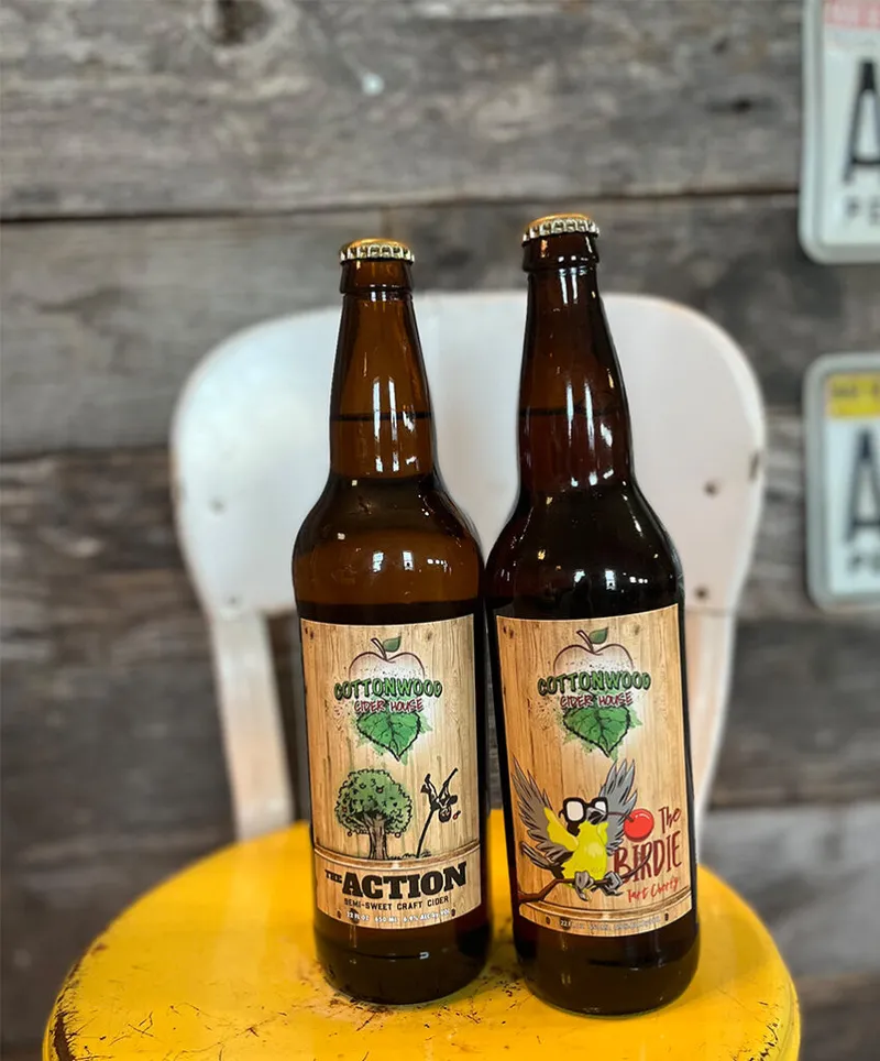 Two Cottonwood Cider House bottles — The Action and The Birdie — on a yellow stool