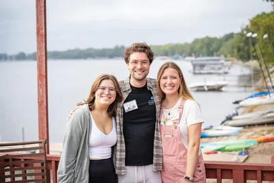 Three smiling young adults posing on a lakeside deck with boats and trees behind them