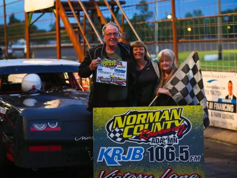 Three people pose in Victory Lane at Norman County Raceway holding a checkered flag and Hobby Stock Feature Winner sign