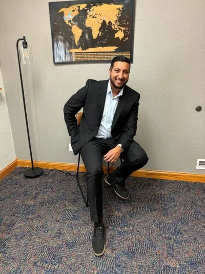 Smiling man in a black blazer seated in an office with a world map on the wall