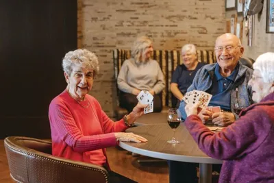 Smiling older adults playing cards at a round table with a glass of red wine nearby