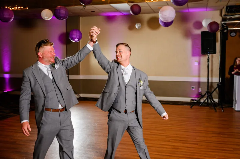 Two grooms in matching gray suits hold hands raised on a dance floor beneath purple paper lanterns