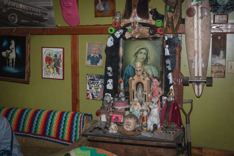 Eclectic wall display with Virgin Mary painting, folk art figures, skateboards, and a rainbow crocheted blanket on a bench