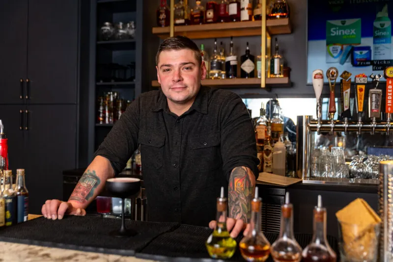 Tattooed bartender leaning on a bar counter with draft taps and liquor bottles behind him