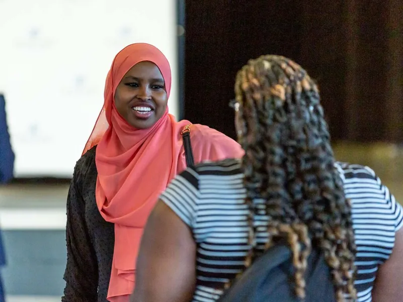 Smiling woman in a coral hijab converses with another woman in a striped top at an indoor event