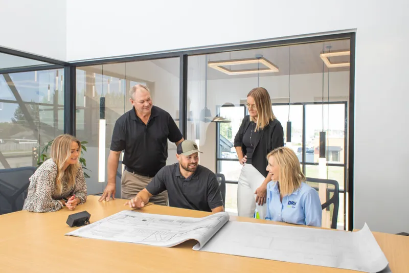 Five colleagues gathered around a conference table reviewing large printed blueprints in a modern glass-walled office