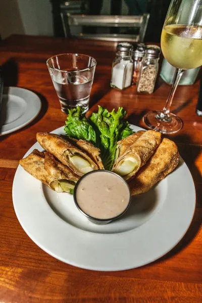 Fried pickle rolls with dipping sauce and lettuce on a white plate beside a glass of white wine