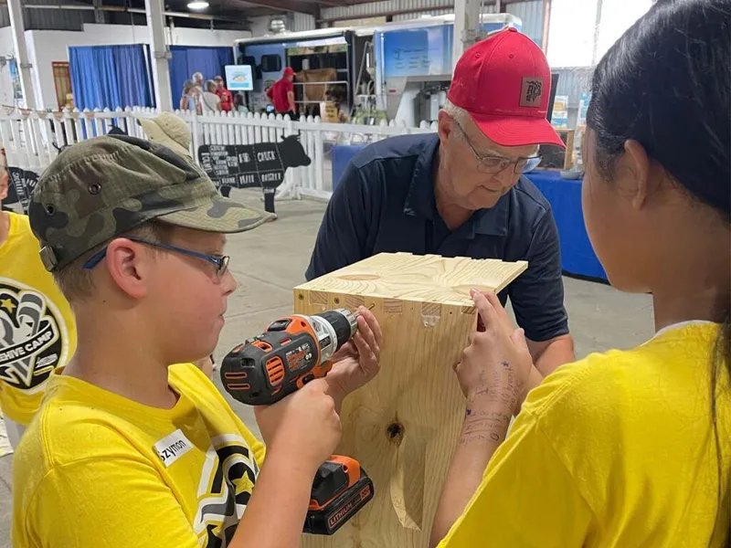 Boy in camo cap uses a power drill on a wood box guided by an adult at a fairground workshop