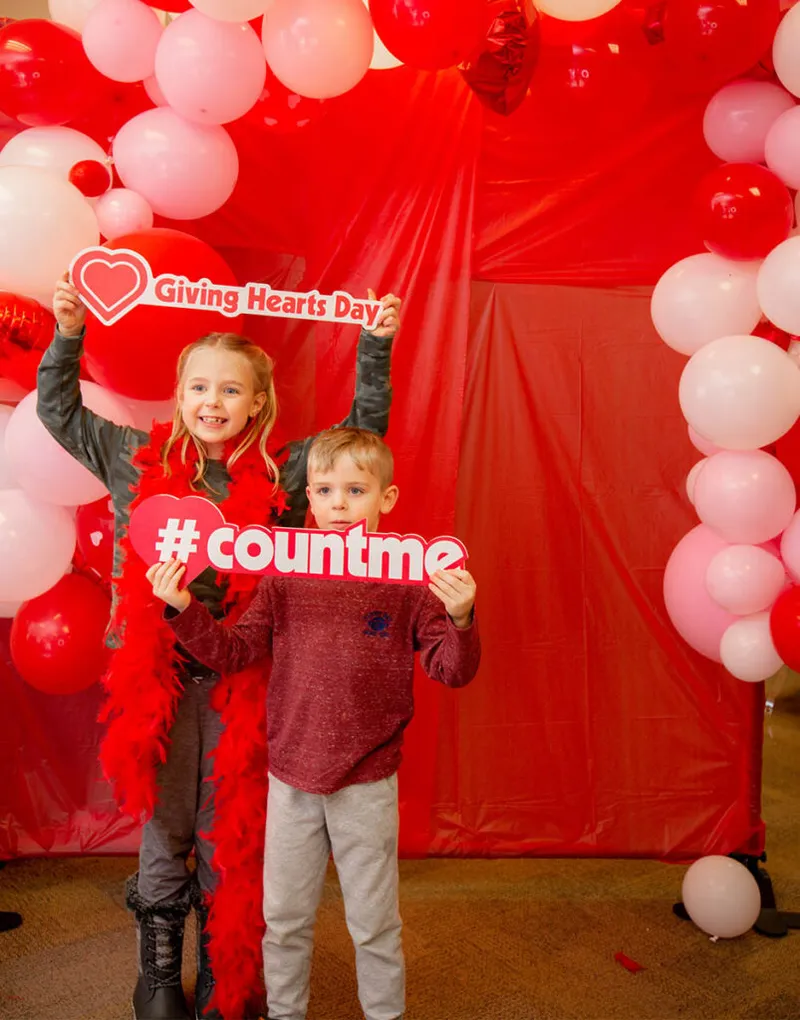 Two children holding Giving Hearts Day and #countme signs in front of a red balloon arch backdrop