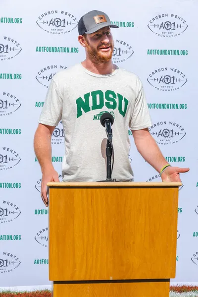 Bearded man in NDSU shirt and cap speaks at podium before AO1 Foundation banner