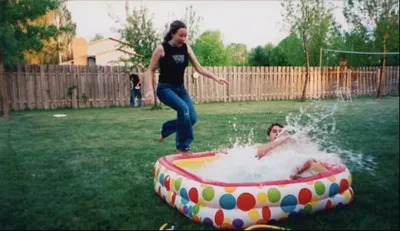 Woman jumping near a colorful inflatable kiddie pool as someone splashes inside, in a fenced backyard