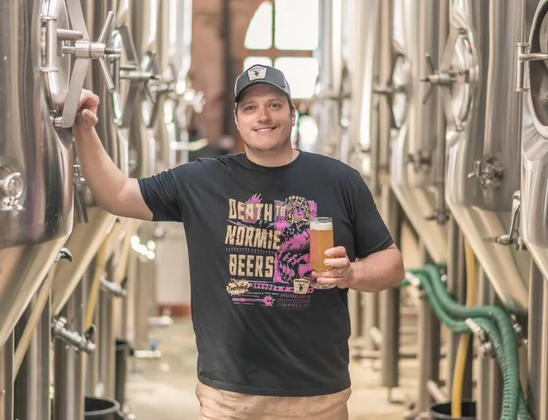 Smiling man holding a pint of beer among stainless steel fermentation tanks in a brewery