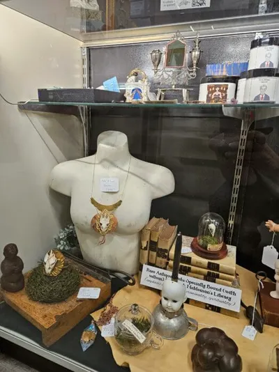 White torso mannequin wearing a statement necklace surrounded by curiosity shop items including skulls, moss, and old books