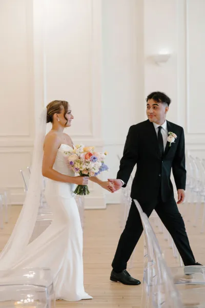 Bride in white gown holding colorful bouquet walks hand-in-hand with groom in black suit down a bright ceremony aisle