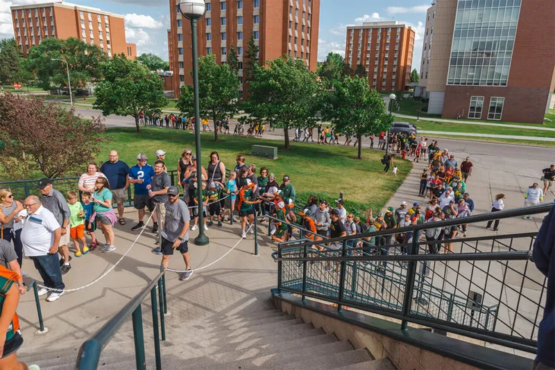 Large crowd queuing along a rope line outside brick campus buildings on a sunny day