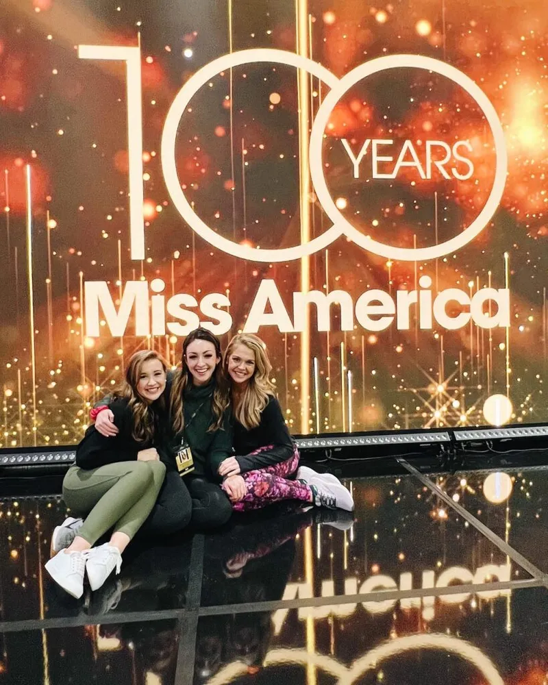 Three young women sitting on a reflective stage in front of a Miss America 100 Years backdrop