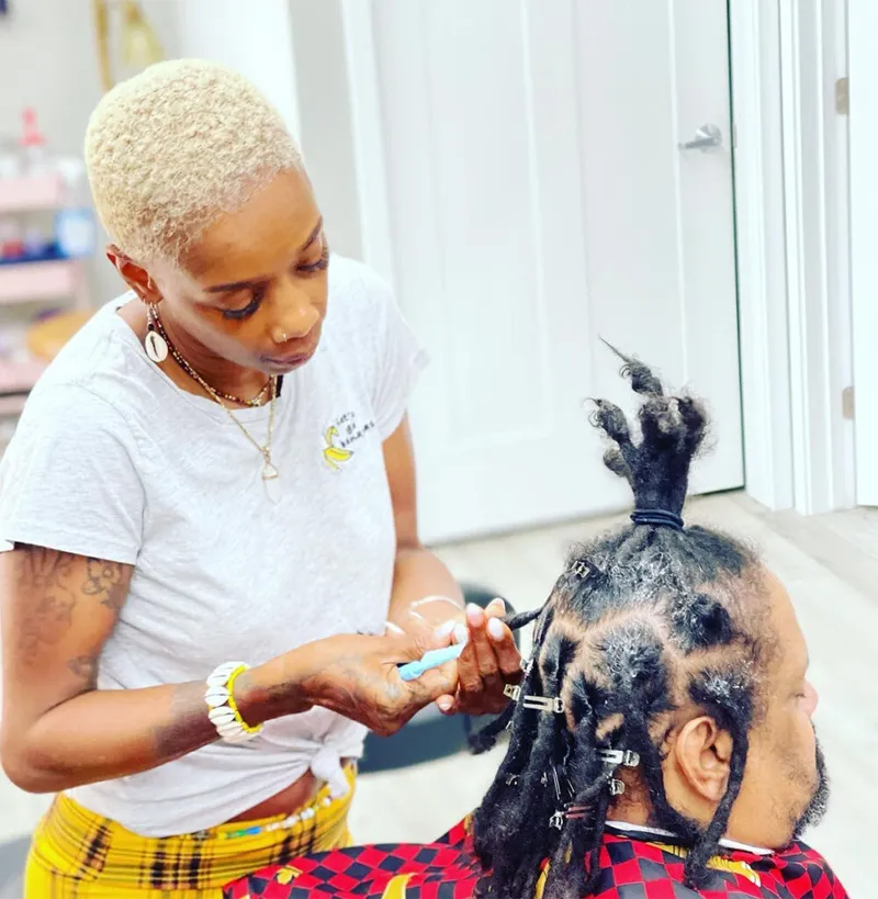 Loctician with short blonde hair applying product to a client's sectioned locs in a bright salon