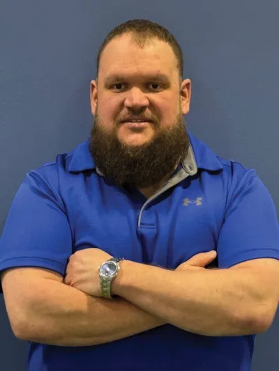 Bearded man in a blue polo shirt standing with arms crossed against a blue wall