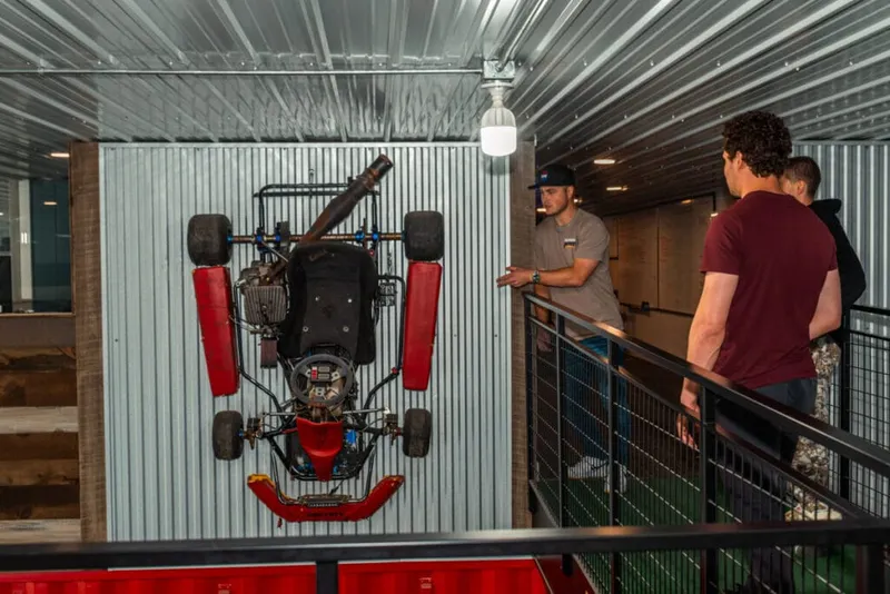 Red go-kart mounted on a corrugated metal wall as two men look on from a mezzanine railing