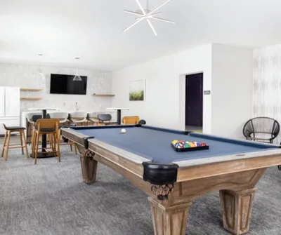Wood-framed pool table with blue felt in a bright modern game room with bar seating and wall-mounted TV