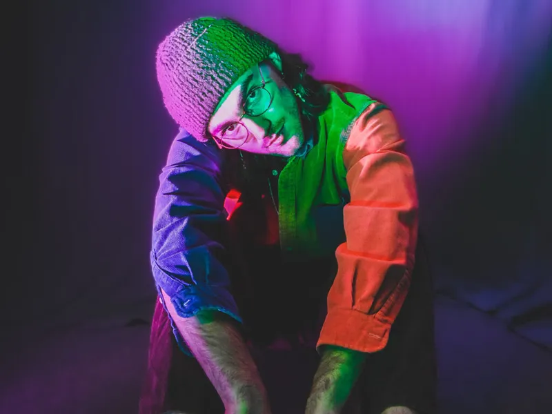 Person in round glasses and knit beanie leaning forward under colorful neon stage lighting