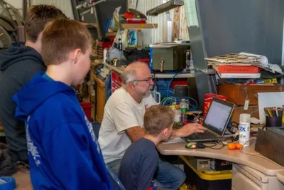 Adult man and two boys gathered around a laptop at a cluttered workshop desk