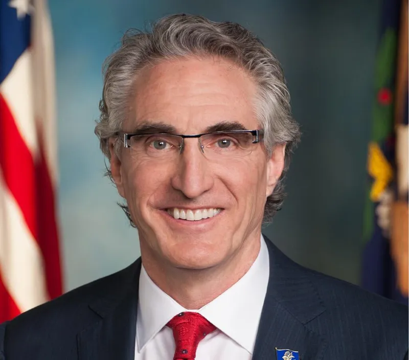 Man with gray hair and glasses in a dark suit and red tie, with US and state flags behind him