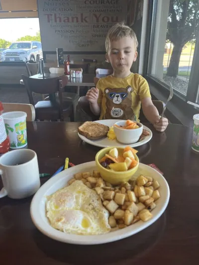 Young boy in a yellow bear shirt at a diner table with breakfast plates of eggs, hash browns, and fruit