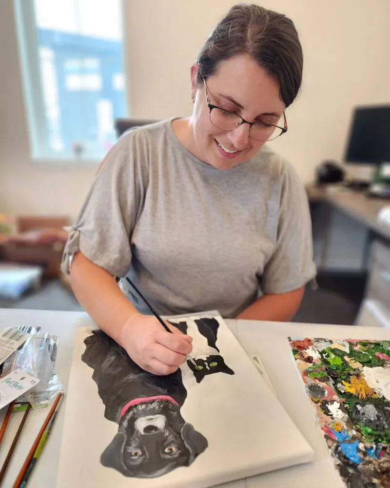 Smiling woman with glasses painting a black dog portrait at a desk with a colorful palette nearby