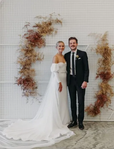 Bride in off-shoulder white gown and groom in black suit pose before a dried floral arch