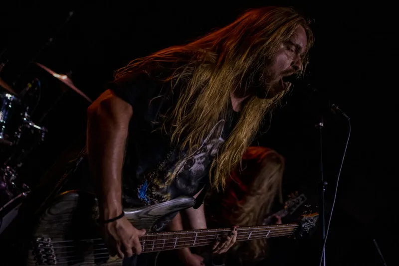Long-haired guitarist screams into a mic while shredding on stage in dark lighting