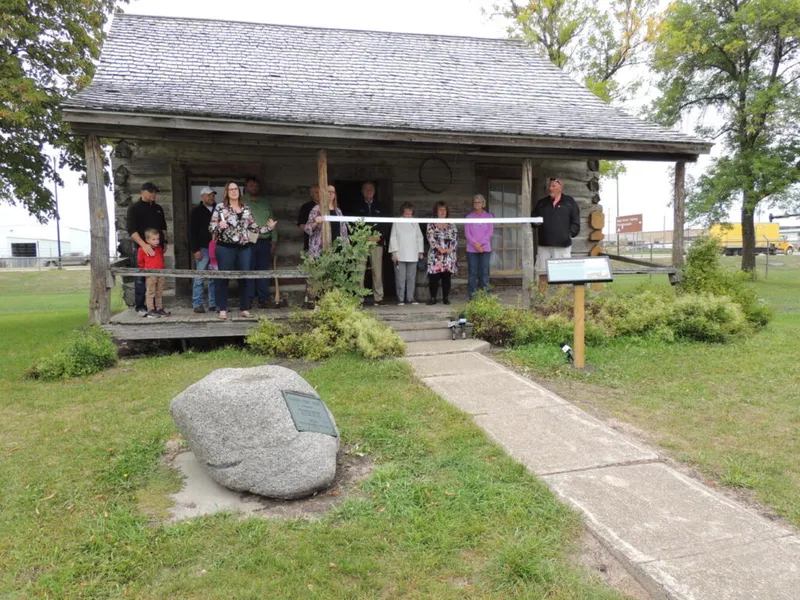 Group of people gathered on the porch of a historic log cabin beside a commemorative boulder and marker