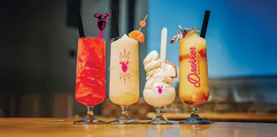Four colorful cocktails in tall glasses garnished with fruit and decorative picks on a wooden bar