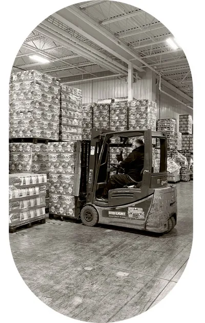 Worker operating a forklift amid tall pallets of canned beer in a large warehouse