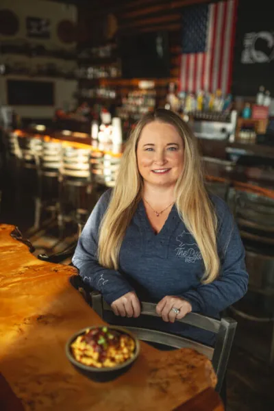 Smiling woman with long blonde hair seated at a wood bar inside a restaurant, with a bowl of mac and cheese in front of her