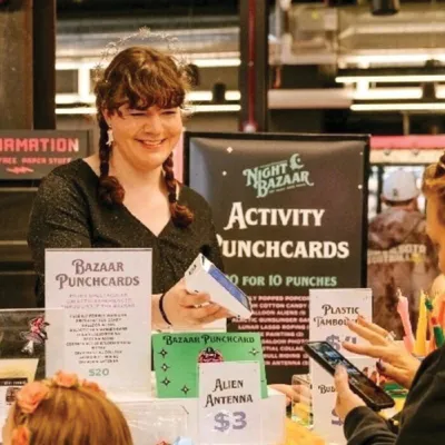 Smiling vendor behind a booth table with Bazaar Punchcards sign and Alien Antenna $3 label at a Night Bazaar event