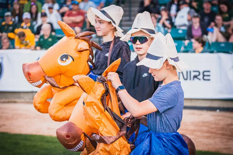 Two kids in bonnet hats race on inflatable horse costumes on a baseball field