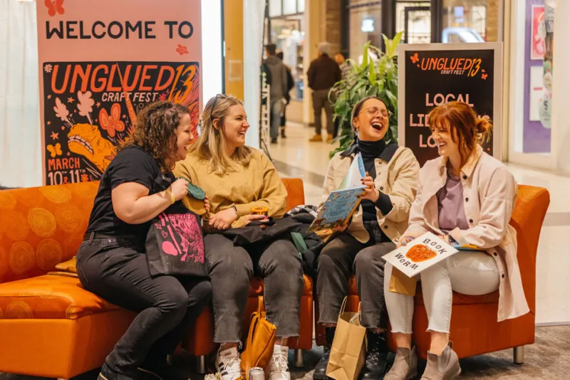 Four women laughing on an orange sofa beside Unglued Craft Fest banners