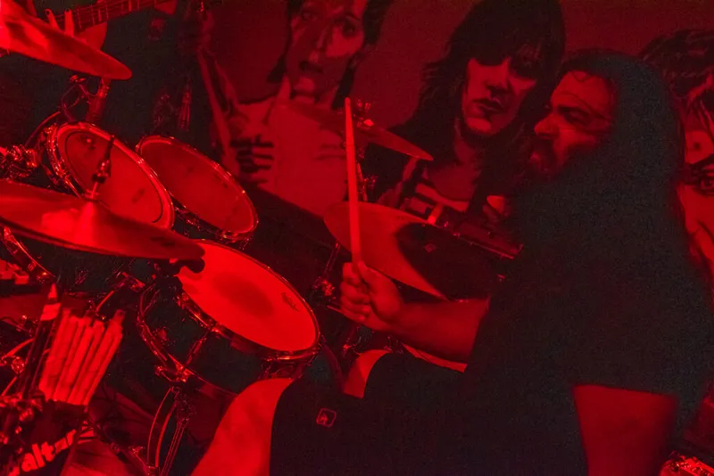 Drummer mid-strike with drumsticks under red stage lighting, bandmates visible behind