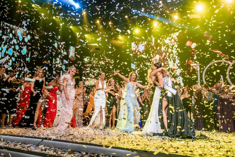 Women in formal gowns celebrate on a lit stage amid a shower of gold and white confetti